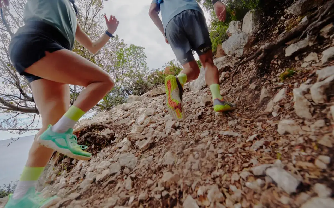 Dwie osoby wbiegają po kamienistym szlaku. Mają na nogach jaskrawe buty 0 drop i kolorowe skarpety.