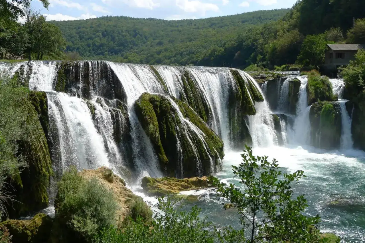 Potężny wodospad na rzece Una, otoczony bujną zielenią. Woda spływa kaskadami po omszałych skałach, tworząc mgiełkę.
