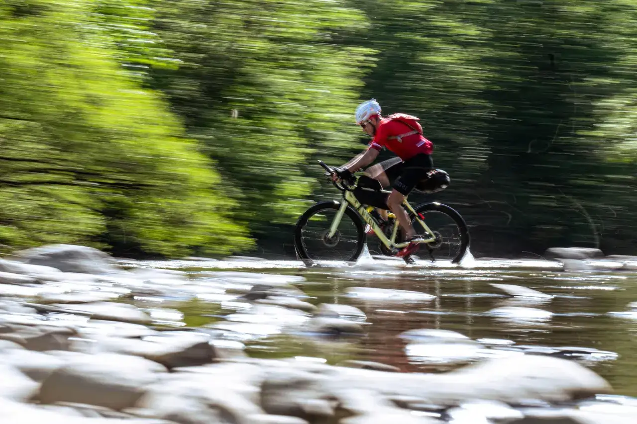 Kolarz w czerwonej koszulce pokonuje wodną przeszkodę podczas ultramaratonu rowerowego.