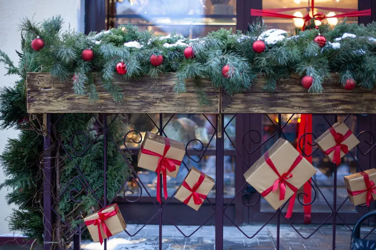Świąteczna girlanda z gałązek iglastych, ozdobiona czerwonymi bombkami i śniegiem, wisi nad płotem z prezentami.