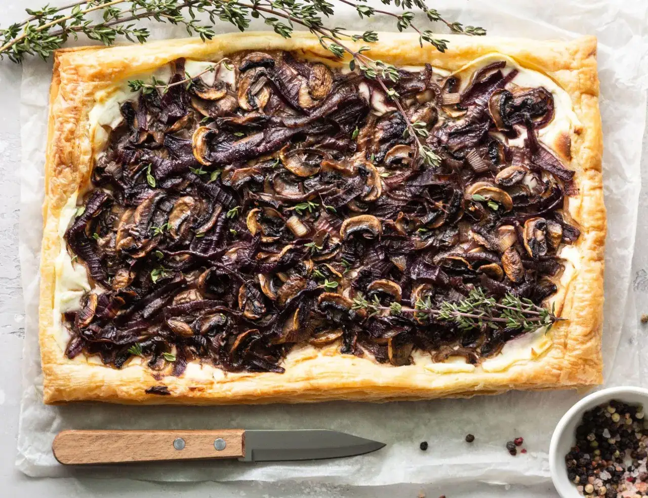 Chrupiące ciasto francuskie z pieczarkami i karmelizowaną cebulą, udekorowane gałązkami tymianku. Idealne na przekąskę.