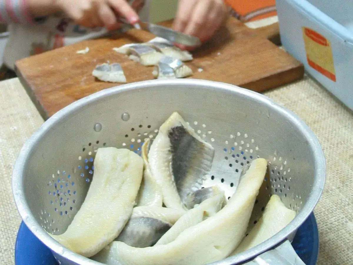 Śledzie w durszlaku czekają na moczenie śledzi w wodzie z octem. W tle krojenie ryby na desce.
