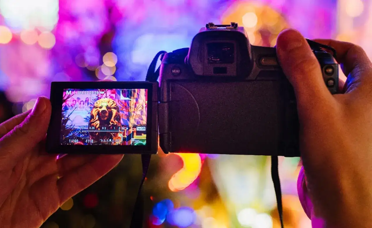 Ręce trzymają aparat Canon EOS, na ekranie widać złoty posąg lwa i kolorowe światła.