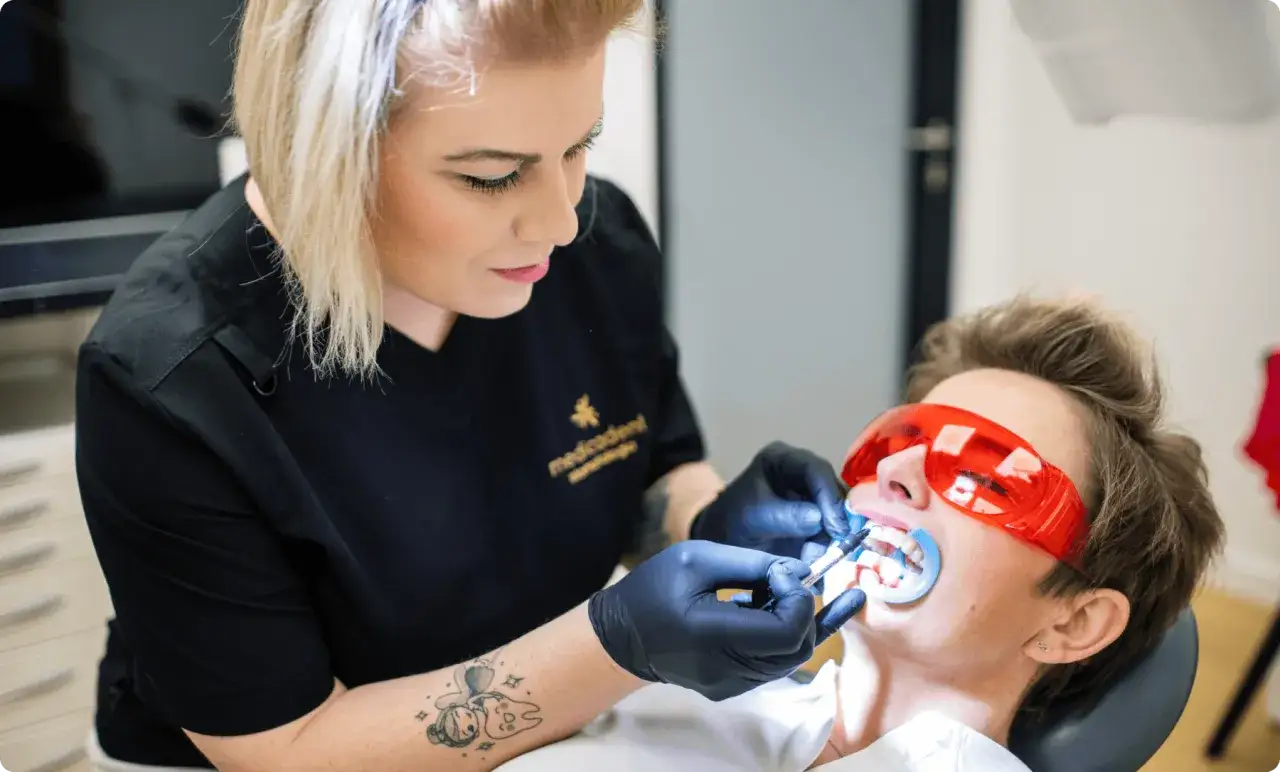 Dentystka wykonuje zabieg wybielania zębów. Pacjentka ma założone okulary ochronne. Czy odkamienianie zębów boli? Nie, to bezbolesny zabieg.