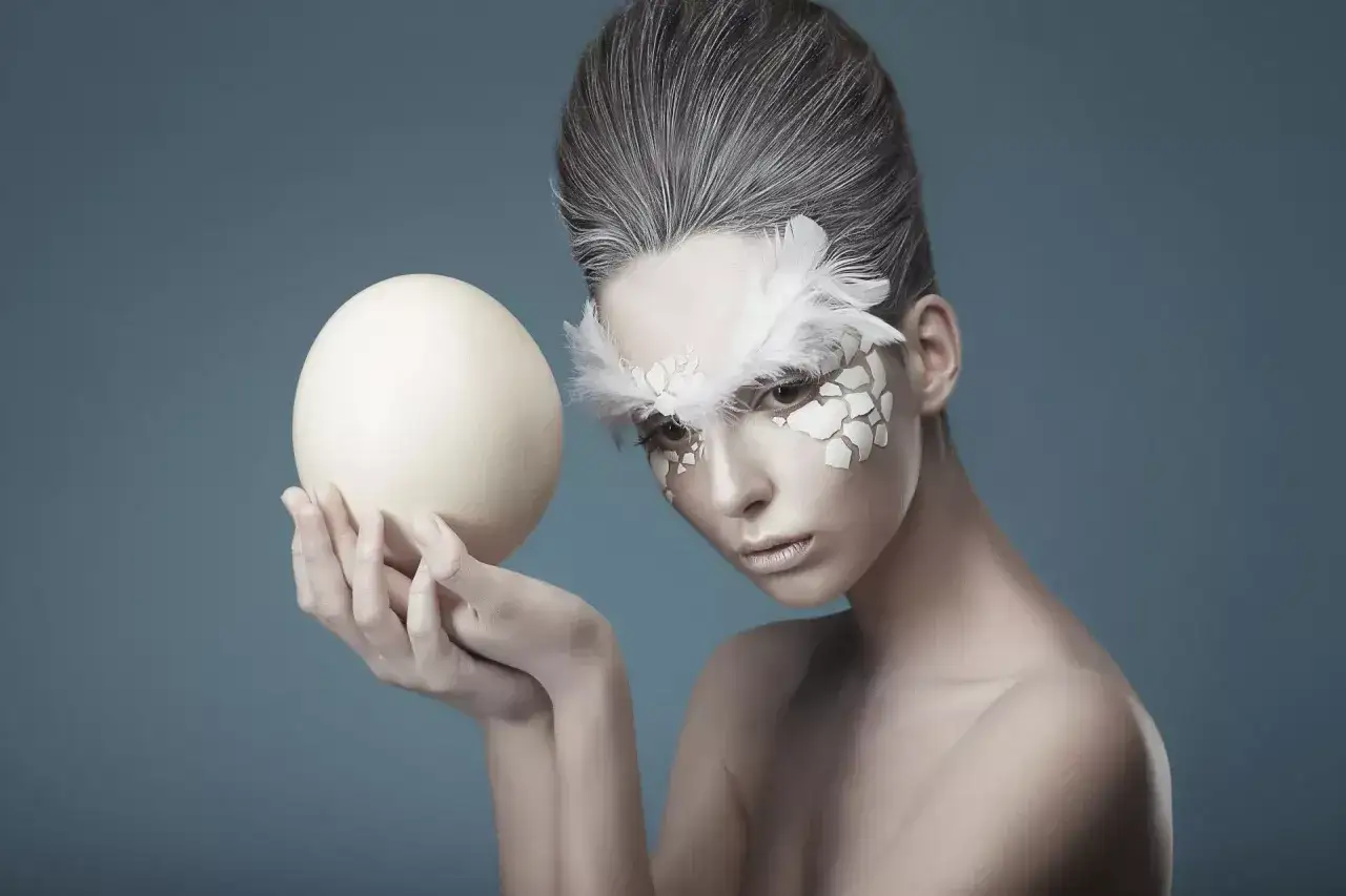 Kobieta z makijażem przypominającym skorupkę jajka i pi&oacute;ra, trzymająca w dłoniach wielkie jajko.