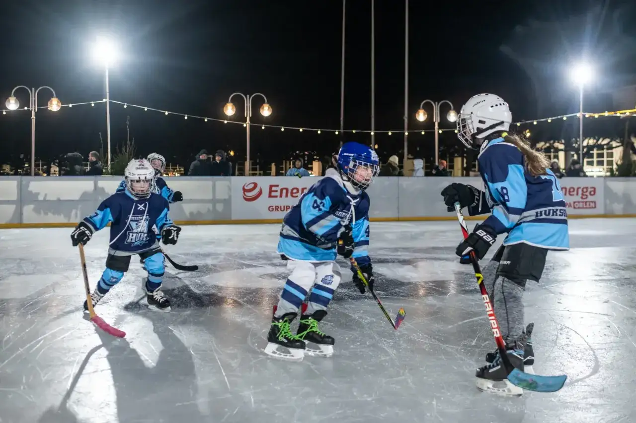 Dzieci w strojach hokejowych ćwiczą dyscyplinę sportową na lodzie. W tle widać widz&oacute;w i oświetlenie nocnego lodowiska.