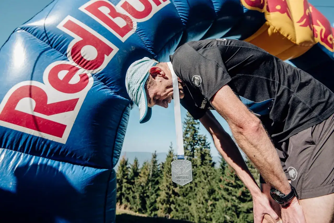 Zmęczony Andrzej Długosz z medalem na szyi pochyla się pod łukiem Red Bulla po ukończonym biegu.