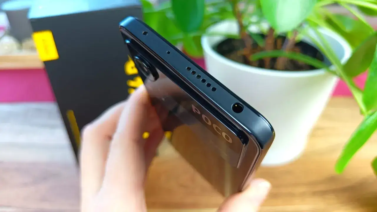 Dłoń trzyma czarny telefon Poco X4, widoczny jest jego bok z głośnikiem i wejściem słuchawkowym.