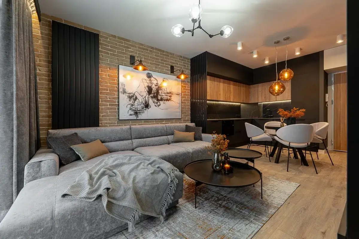 Nowoczesne, małe mieszkanie z szarą sofą, ceglaną ścianą i aneksem kuchennym.