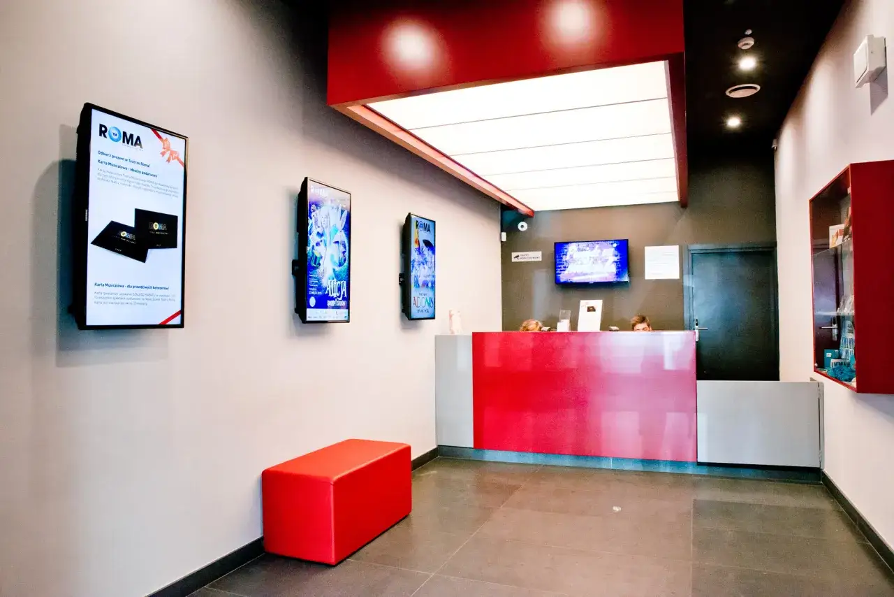 W holu Teatru Muzycznego Roma Nowogrodzka Warszawa, czerwona lada i ławka, plakaty spektakli na ścianach.
