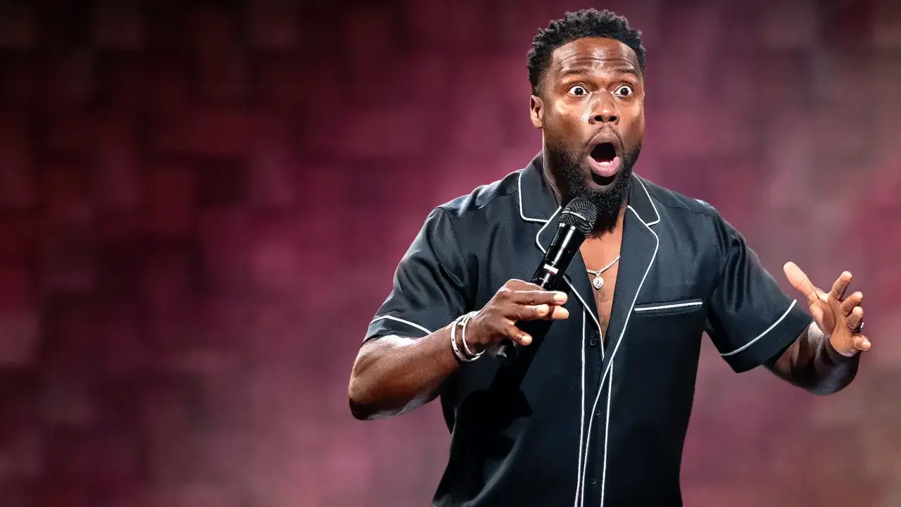 Kevin Hart stand up na scenie, z mikrofonem w dłoni, z zaskoczoną miną i rozłożonymi rękami.