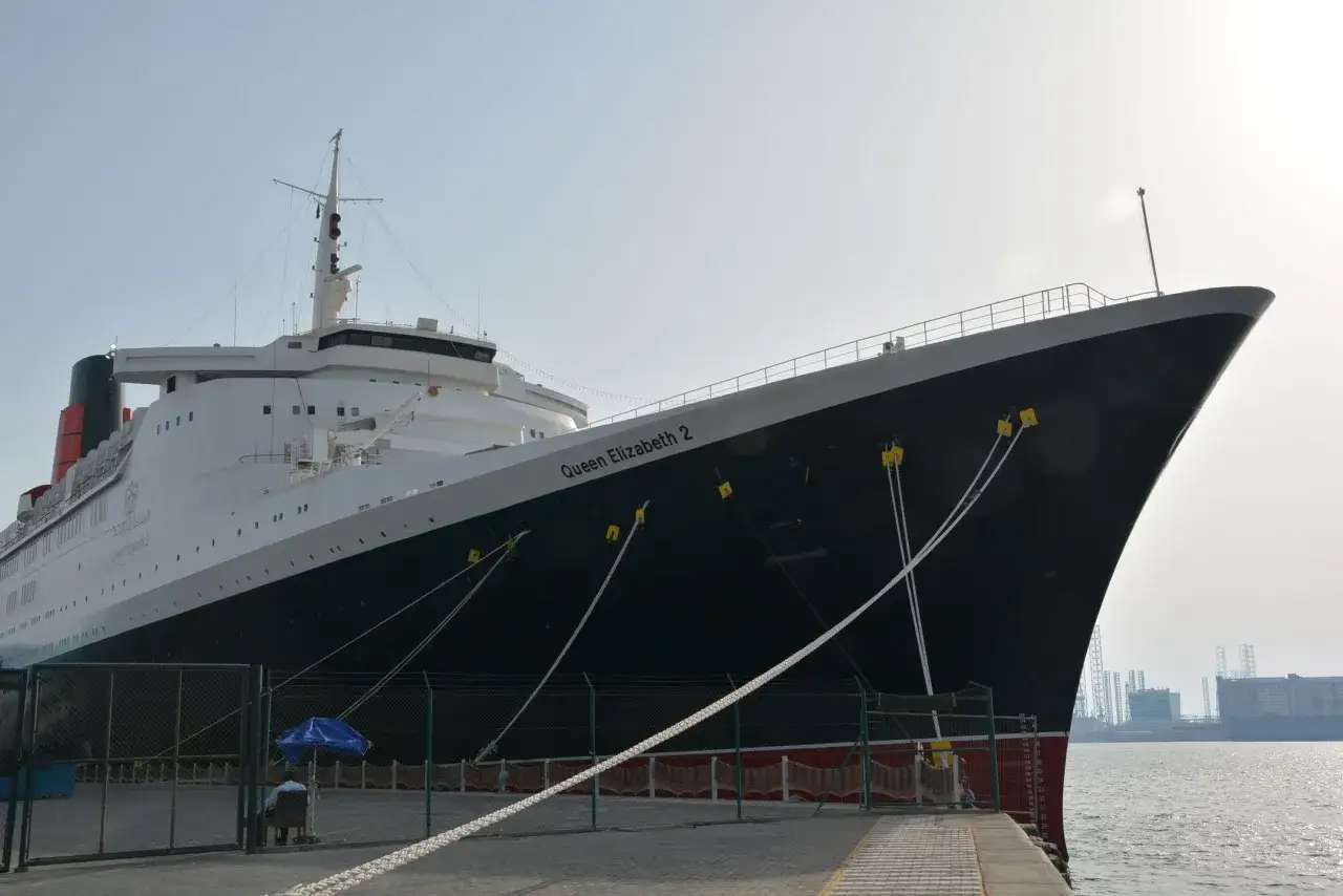 Ogromny statek Queen Elizabeth 2 cumuje przy nabrzeżu. W tle widać przemysłowy krajobraz.