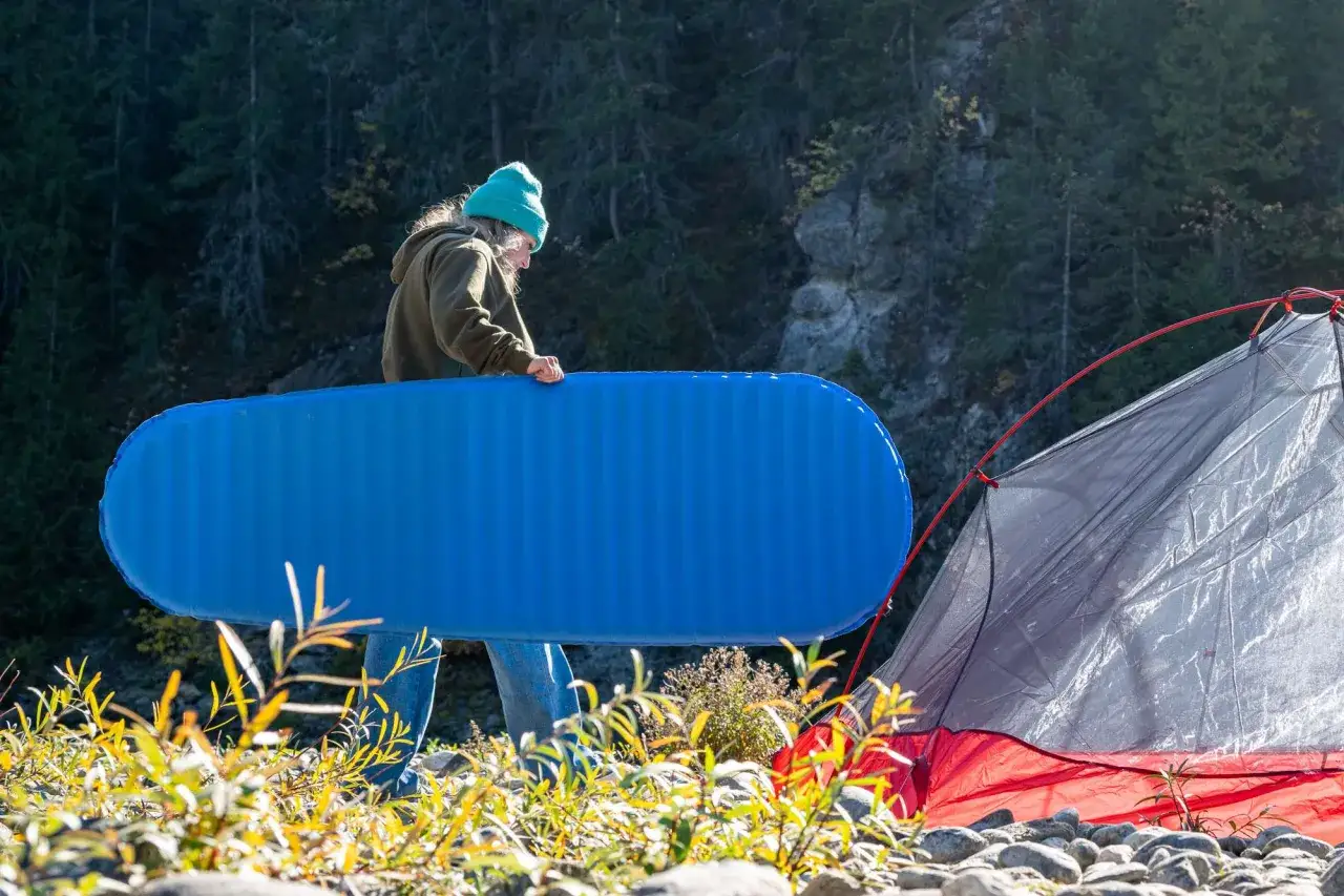 Osoba w czapce przygotowuje niebieski materac, jeden z niezbędnych gadżet&oacute;w pod namiot, obok rozstawionego namiotu.
