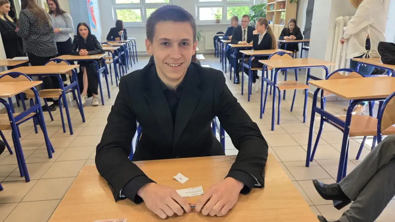 Uśmiechnięty uczeń w garniturze siedzi w sali lekcyjnej. To ważny moment w jego drodze do wykształcenia w Polsce.