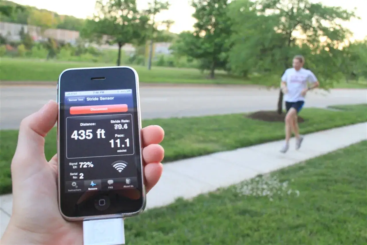 Dłoń trzyma iPhone'a z aplikacją do biegania, pokazującą dystans 435 st&oacute;p i tempo 11.1 min/milę. W tle biegnie mężczyzna. Ant+ co to? To technologia łączności.