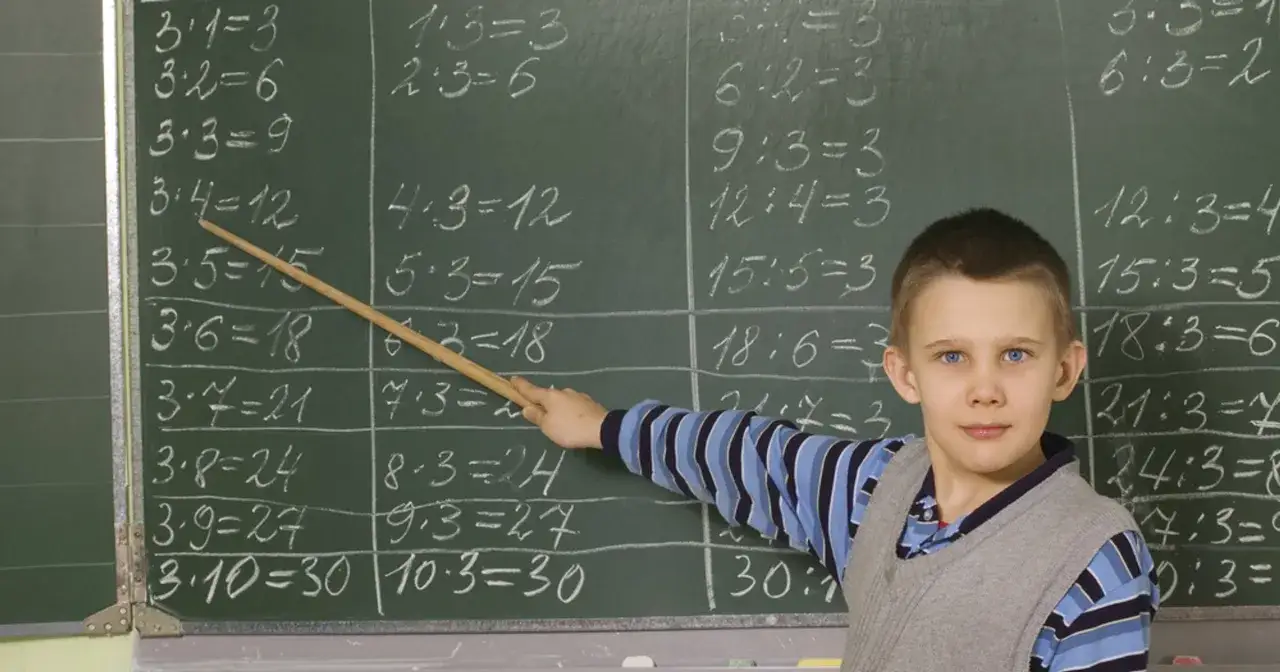 Chłopiec z niebieskimi oczami wskazuje patykiem na tablicę z zapisaną tabliczką mnożenia do 10.