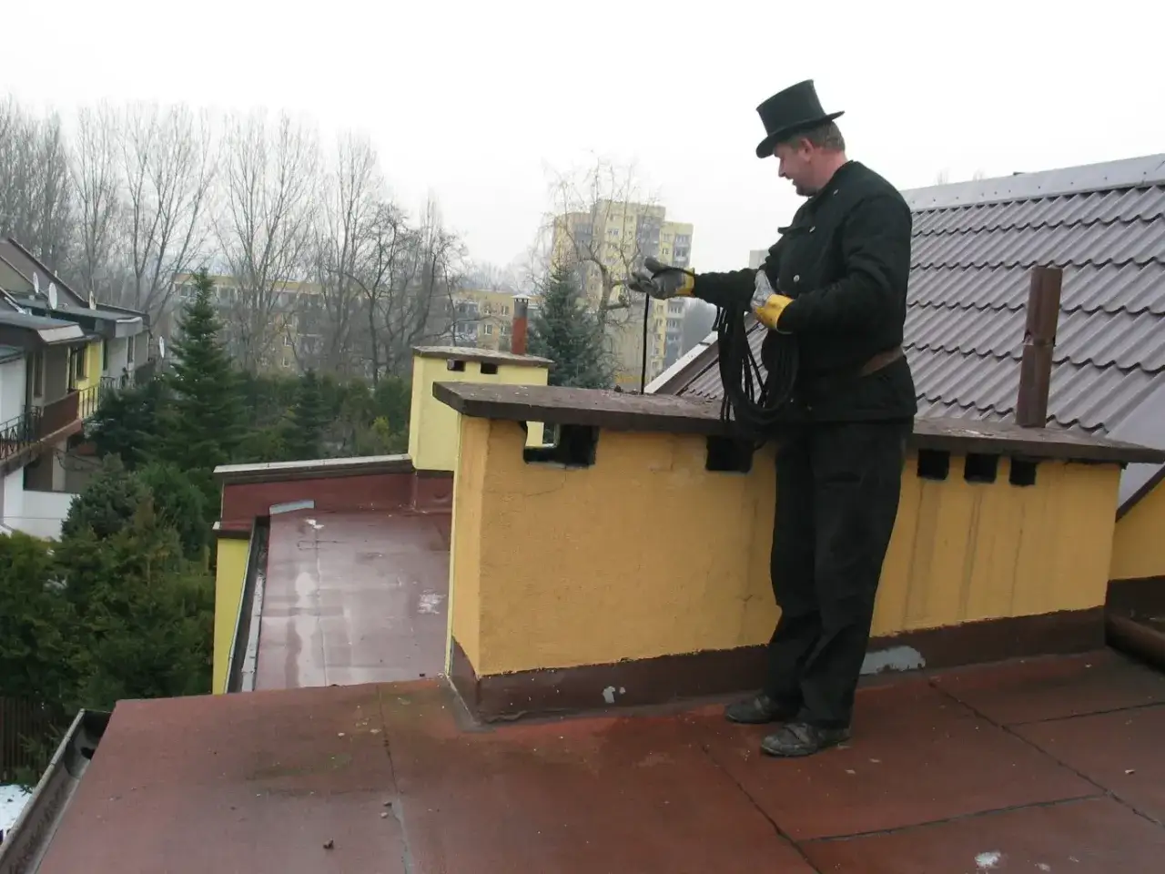 Kominiarz w cylindrze na dachu wykonuje przegląd kominiarski w mieszkaniu bez gazu.