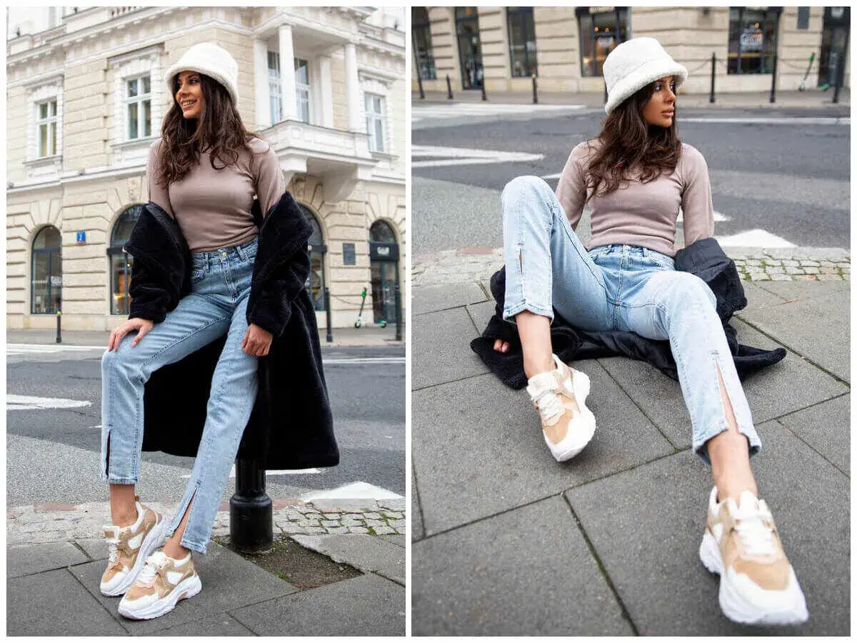 Kobieta w jasnych jeansach z rozcięciami, beżowym sweterku i białej czapce typu bucket hat, siedzi na chodniku.