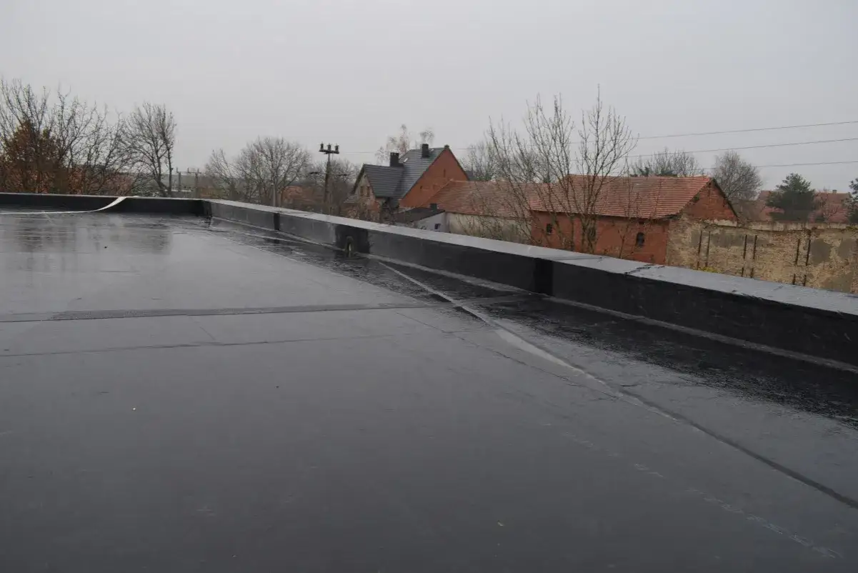 Czarna, błyszcząca powierzchnia dachu płaskiego, wykonana z materiału EPDM. W tle widać budynki i drzewa.