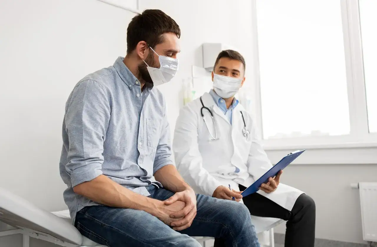 Lekarz w masce i białym fartuchu rozmawia z pacjentem o tym, jak wygląda badanie urologiczne u mężczyzn.