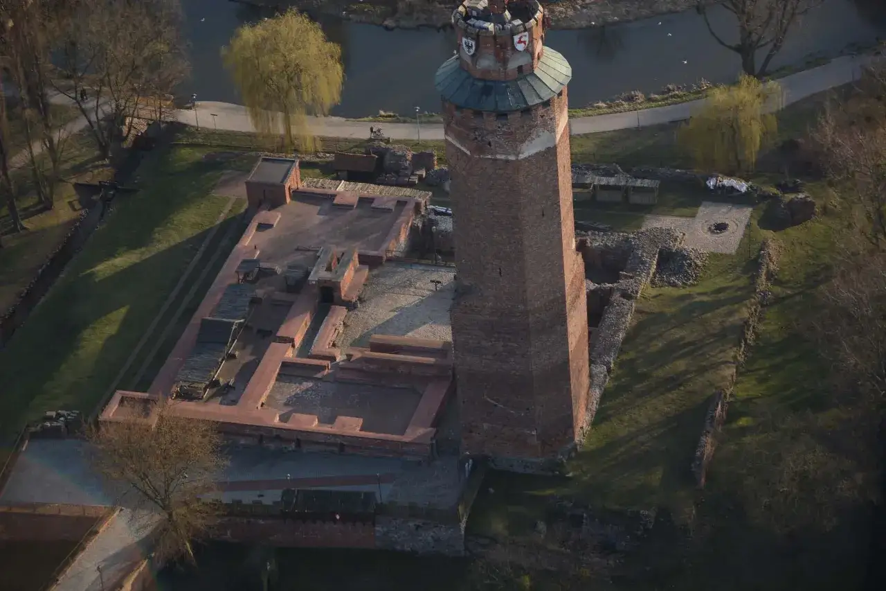 Zamek krzyżacki w Brodnicy, potężna wieża i ruiny dziedzińca, otoczone zielenią i wodą.