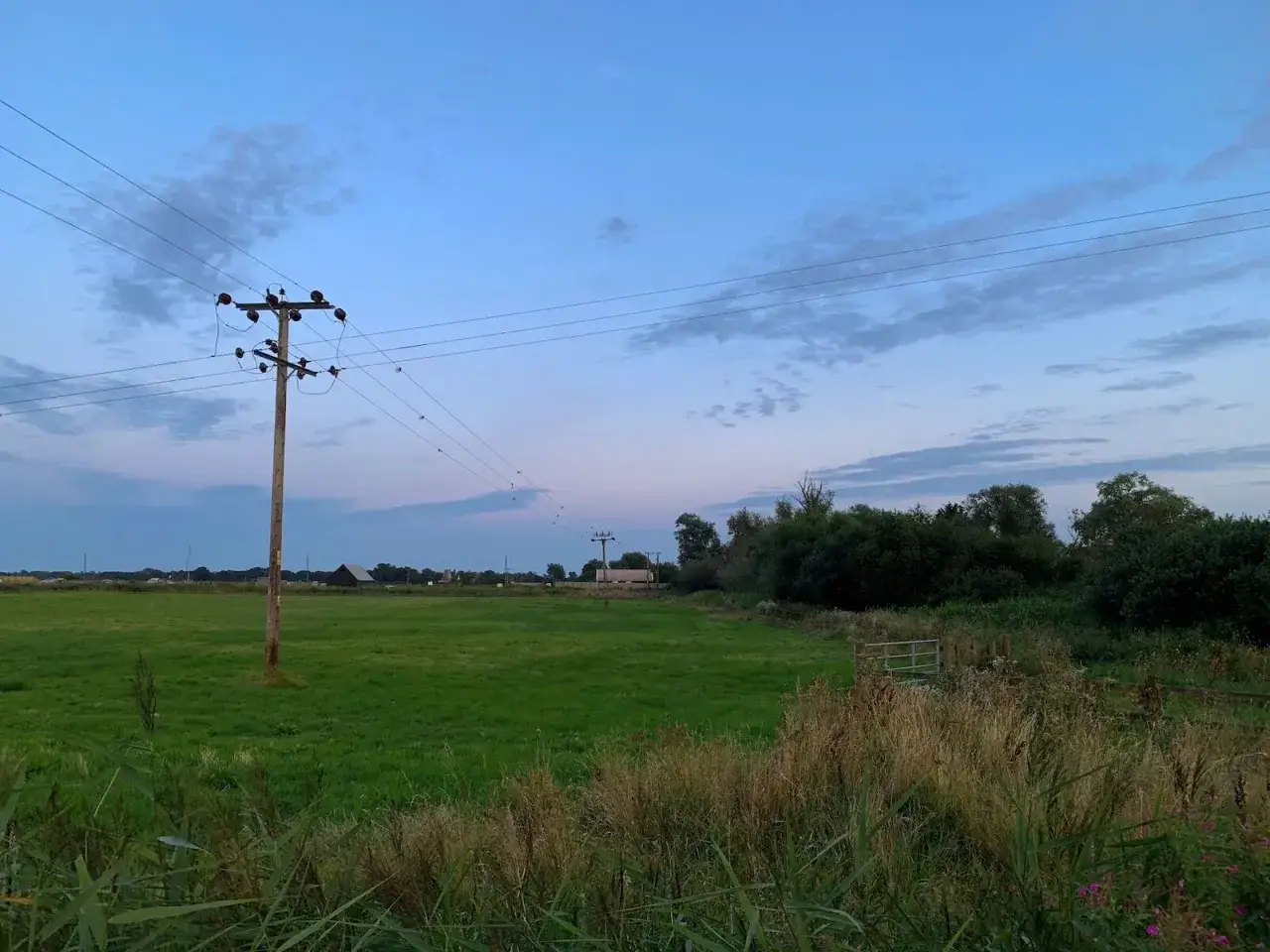 Słup energetyczny stoi na zielonej łące, widać jego odległość od granicy działki i linii energetycznej biegnącej przez krajobraz.