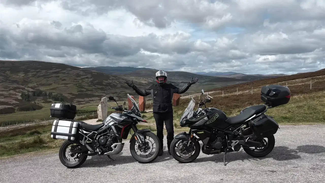 Motocyklista w kasku i kurtce pozuje obok dw&oacute;ch motocykli: szarego i czarnego **Tiger 900 GT Pro**. W tle malowniczy krajobraz g&oacute;rski.