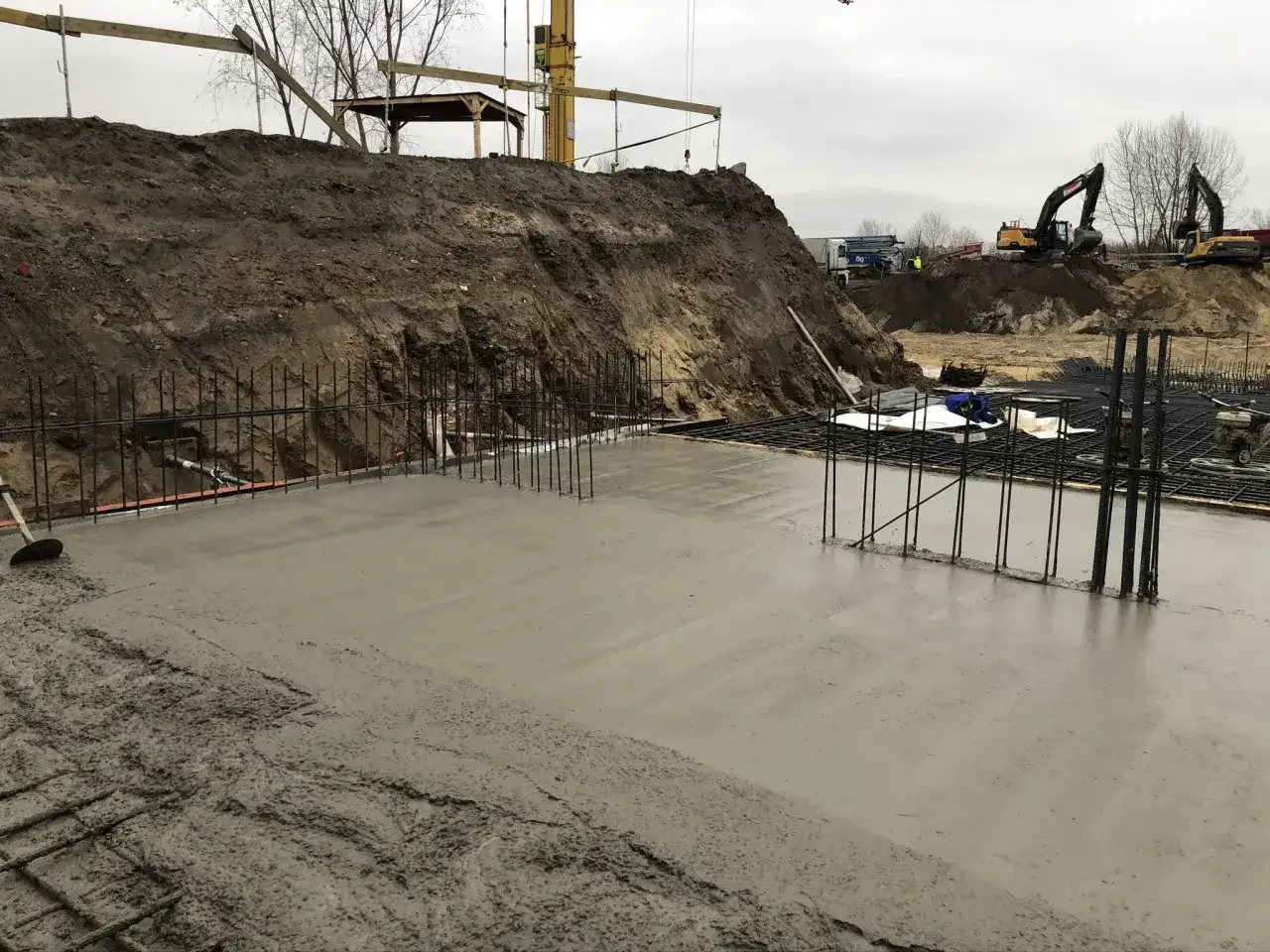 Budowa fundamentów: świeża wylewka betonowa, zbrojenie i koparki w tle. Jak zrobić wylewkę betonową? Tu widać początek prac.