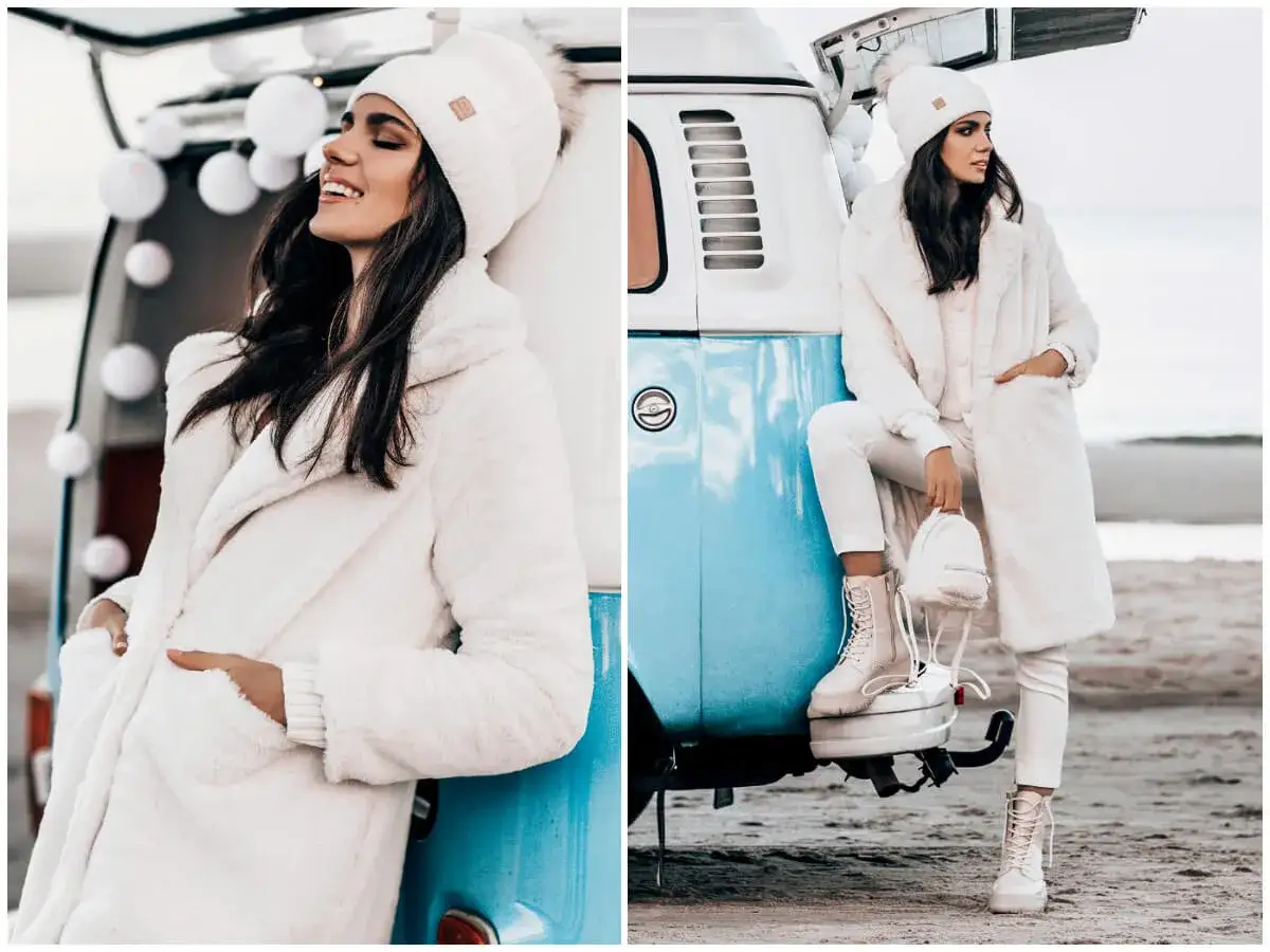 Kobieta w białym, puchatym płaszczu i czapce z pomponem prezentuje idealny outfit na zimę. Ubrana w białe spodnie i trapery, pozuje przy turkusowym vanie na plaży.