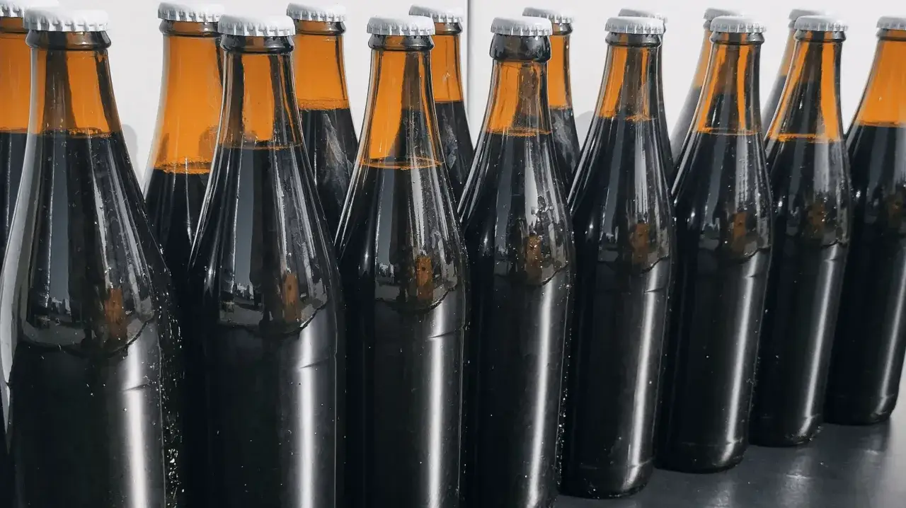 Szereg ciemnych butelek z piwem, każda z białym kapslem. Zastanawiasz się, ile może stać otwarte piwo? Te wyglądają na świeżo zamknięte.