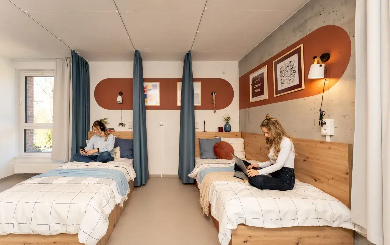 Dwie dziewczyny w nowoczesnym pokoju, przypominającym hostel lub hotel. Jedna korzysta z telefonu, druga z laptopa.