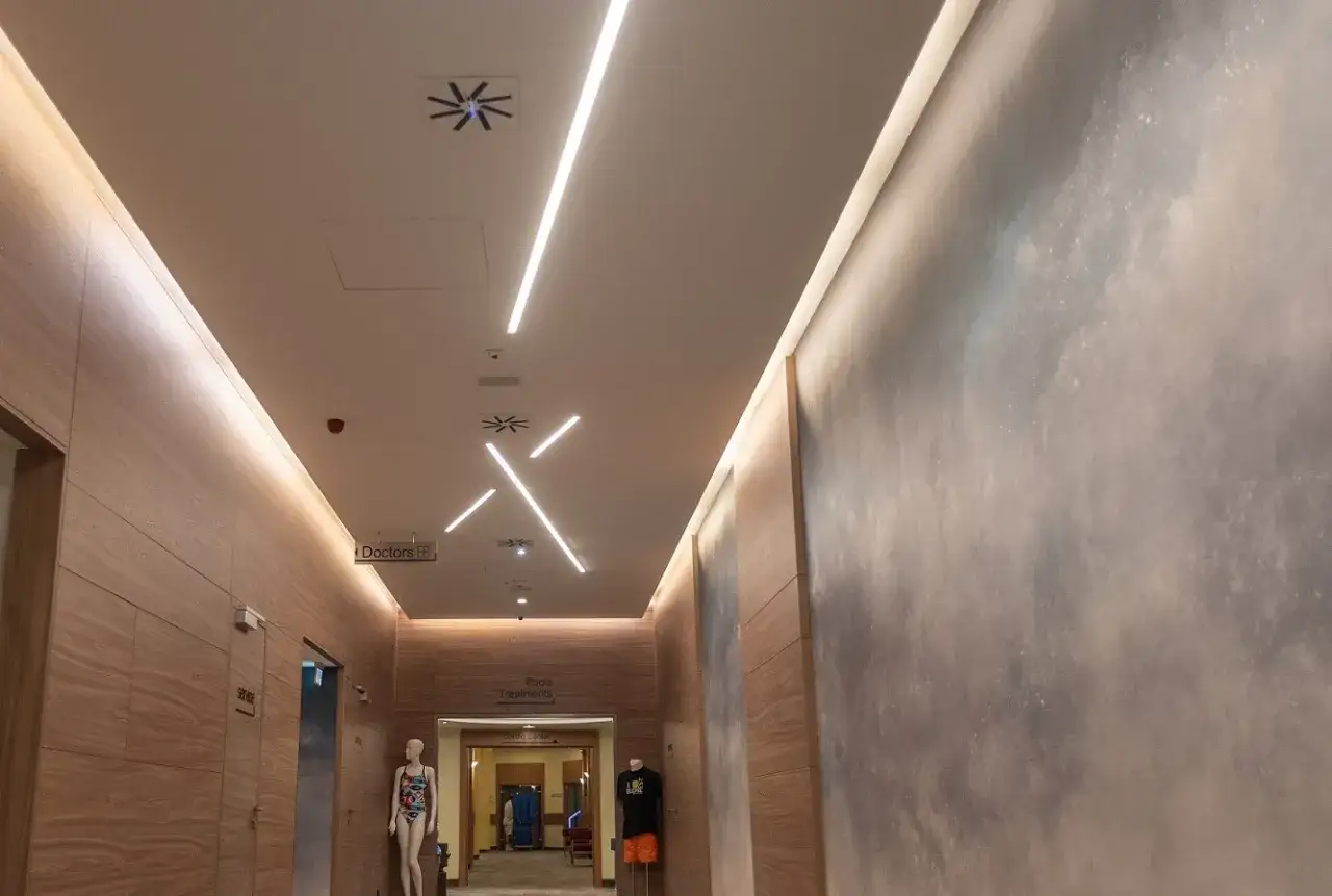 Nowoczesne ledy na suficie bez podwieszanego sufitu oświetlają korytarz z drewnianymi panelami. Widać znaki 