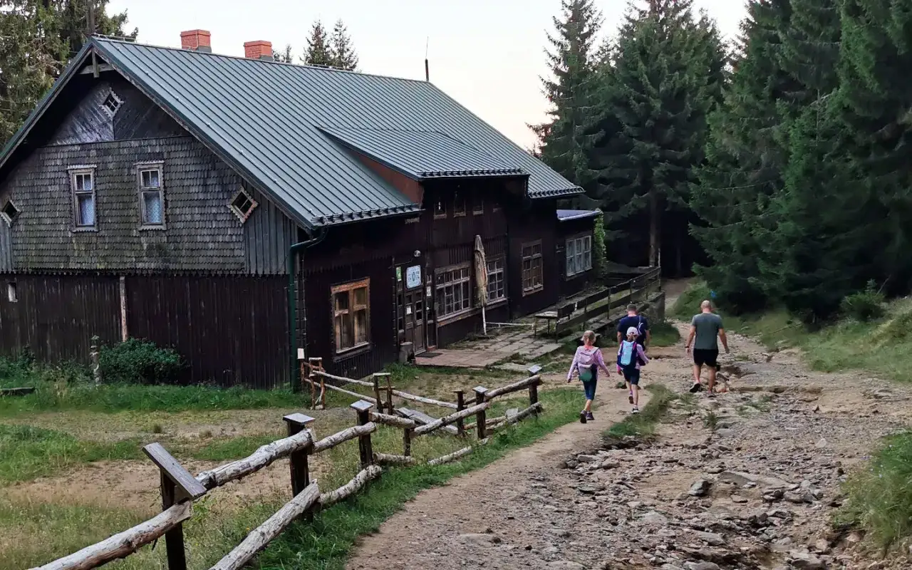 Rodzina idzie ścieżką do schroniska w górach. Drewniany budynek z charakterystycznym dachem otoczony jest lasem.
