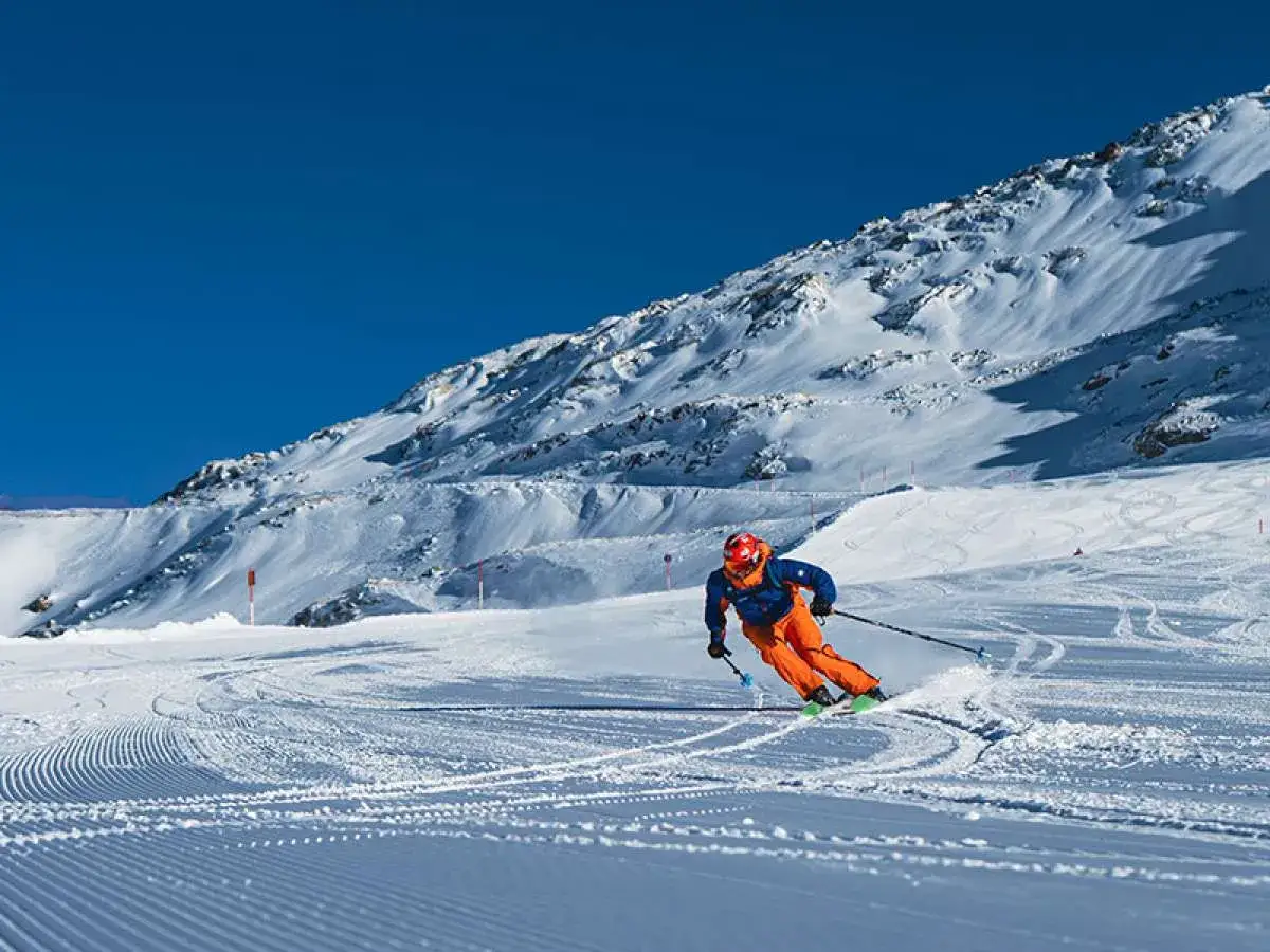 Narciarz w pomarańczowych spodniach i niebieskiej kurtce zjeżdża po ośnieżonym stoku. Posiadanie skipassu to klucz do tej zimowej przygody.