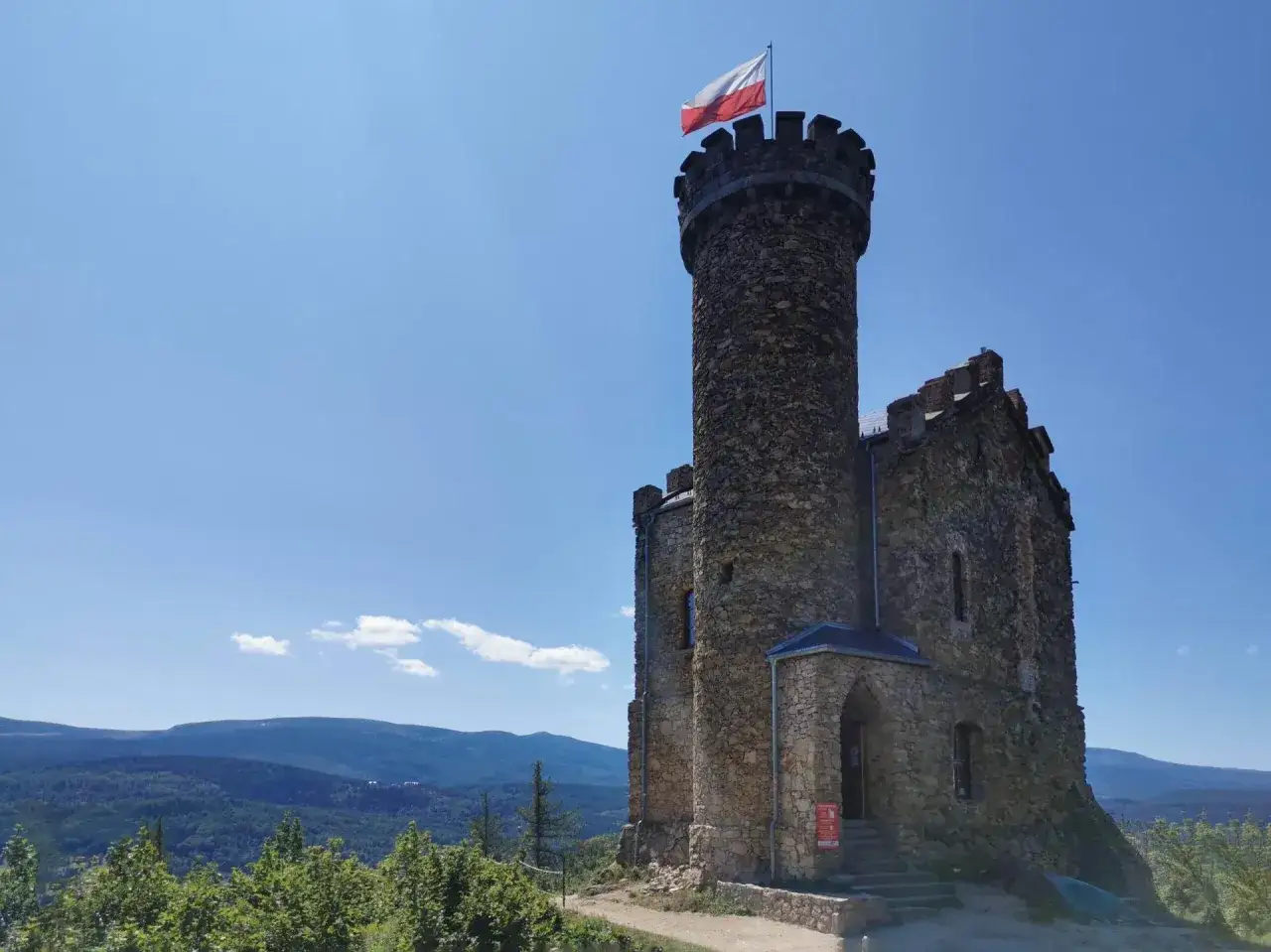 Kamienny zamek księcia Henryka na trasie widokowej, z polską flagą na wieży. W tle góry i lasy.