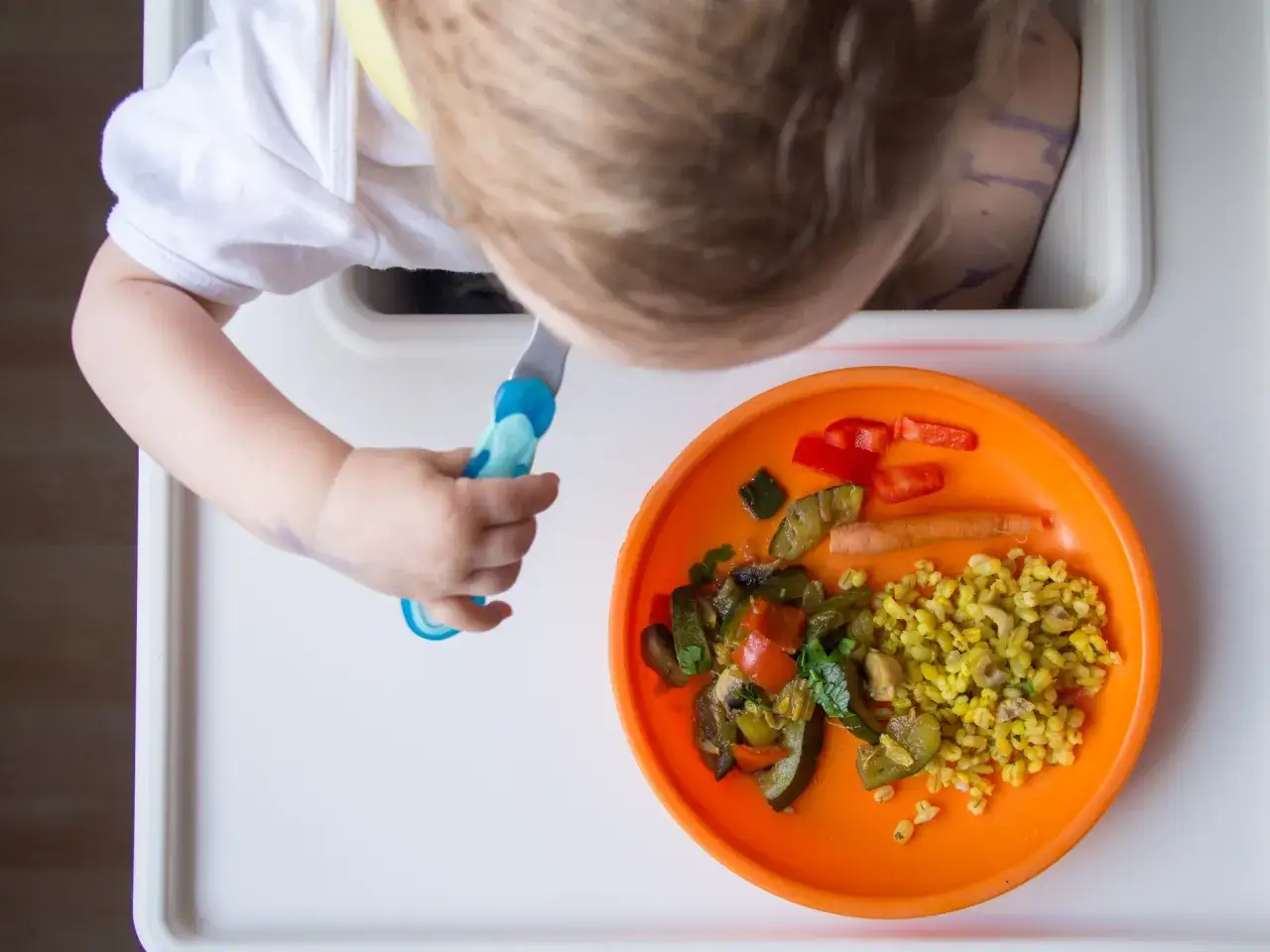 Małe dziecko próbuje samodzielnie jeść obiadki dla 6 miesięcznego dziecka: kaszę z warzywami i kawałki papryki.