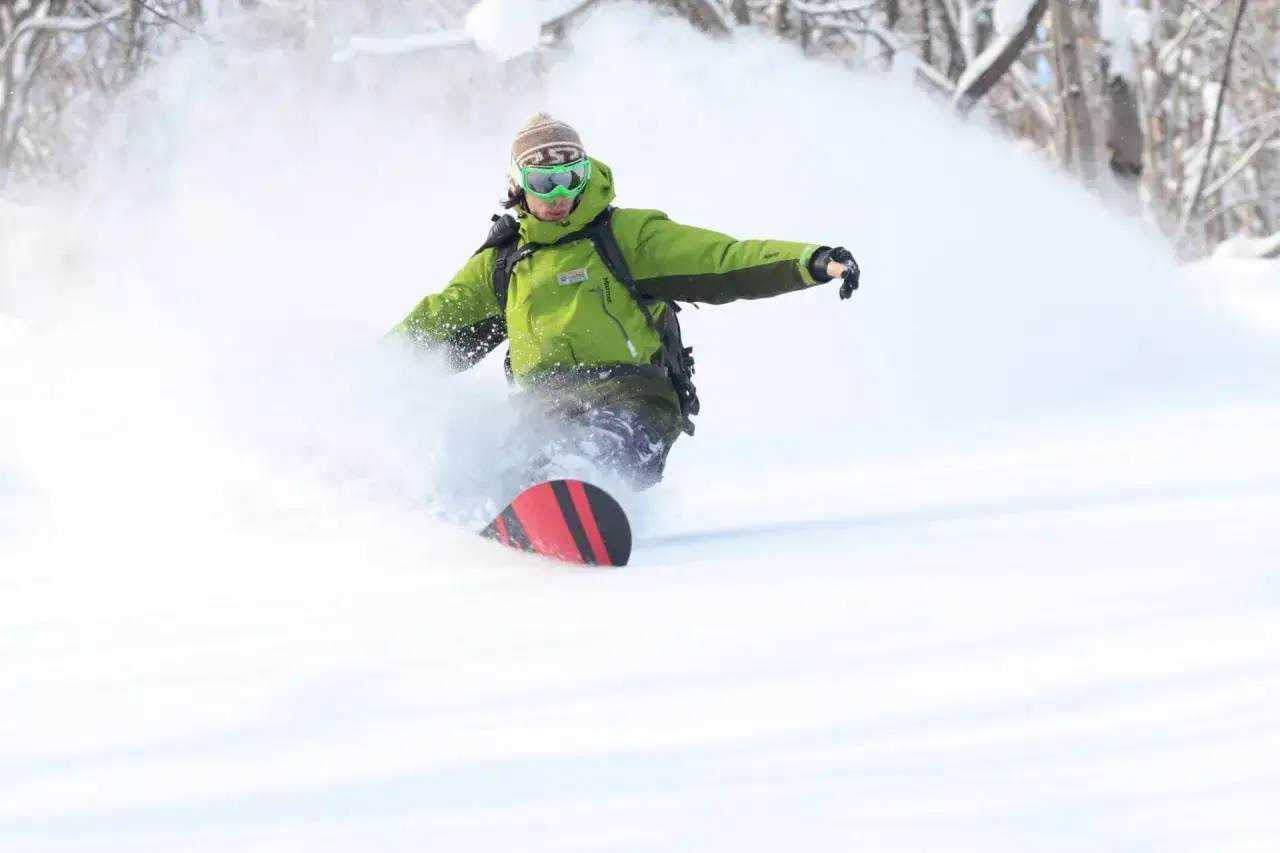 Snowboardzista w zielonej kurtce i goglach szaleje na śniegu, tworząc obłoki puchu. Jego deski do freeride z czerwonymi pasami tną białą przestrzeń.