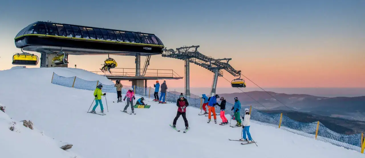 Grupa narciarzy i snowboardzistów na ośnieżonych stokach góry, pod nowoczesną kolejką linową. Zachód słońca maluje niebo na pastelowe kolory.