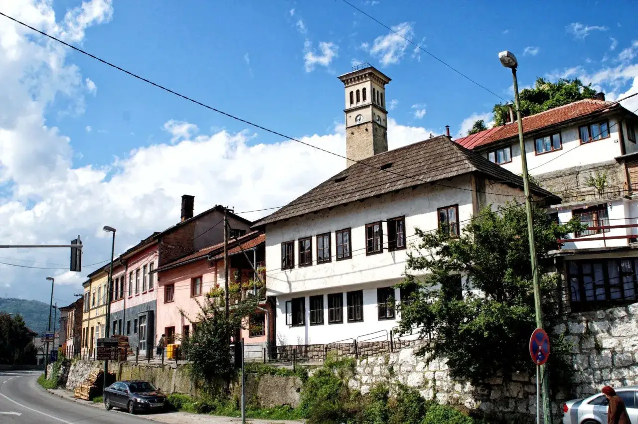 Wieża zegarowa g&oacute;ruje nad kolorowymi domami w **Travnik, Bośnia**. Słoneczny dzień, chmury na niebie.