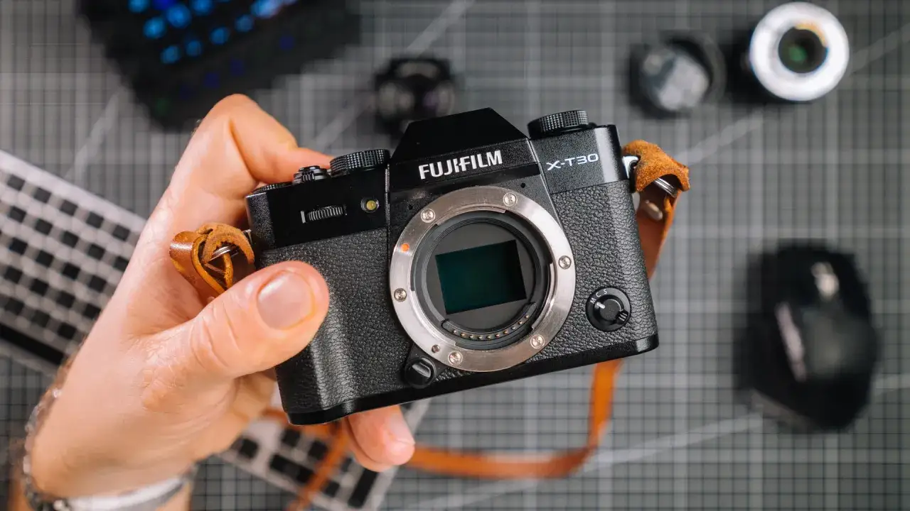 Dłoń trzyma aparat Fujifilm X-T30, który aparat fotograficzny wybrać? W tle widać akcesoria fotograficzne.