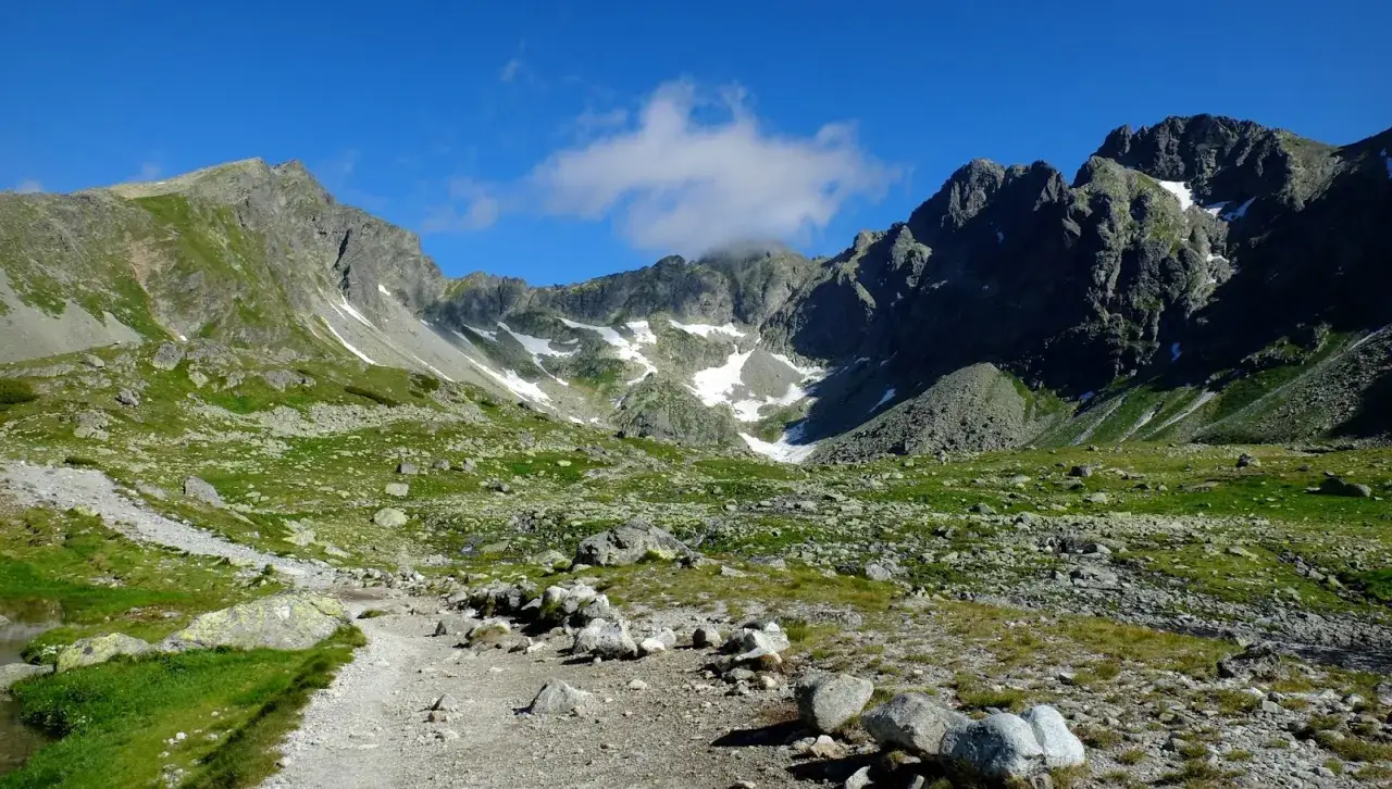 Kamienista ścieżka prowadzi przez zieloną dolinę ku skalistym zboczom. W oddali widać ośnieżony szczyt w Tatrach Wysokich, a nad nim błękitne niebo z pojedynczą chmurką.