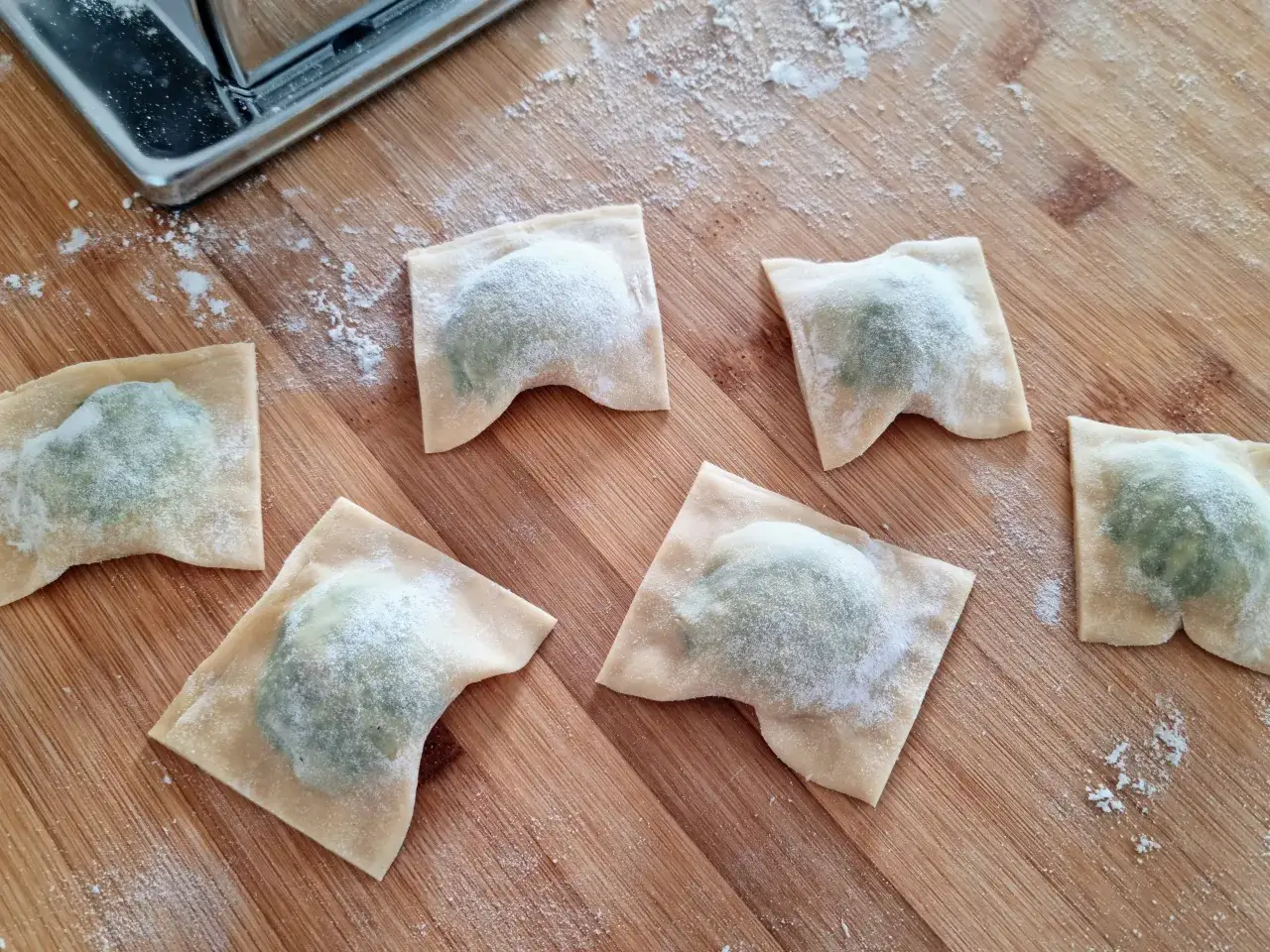 Sześć świeżych ravioli z ricottą, posypanych mąką, leży na drewnianej desce obok wałkownicy.