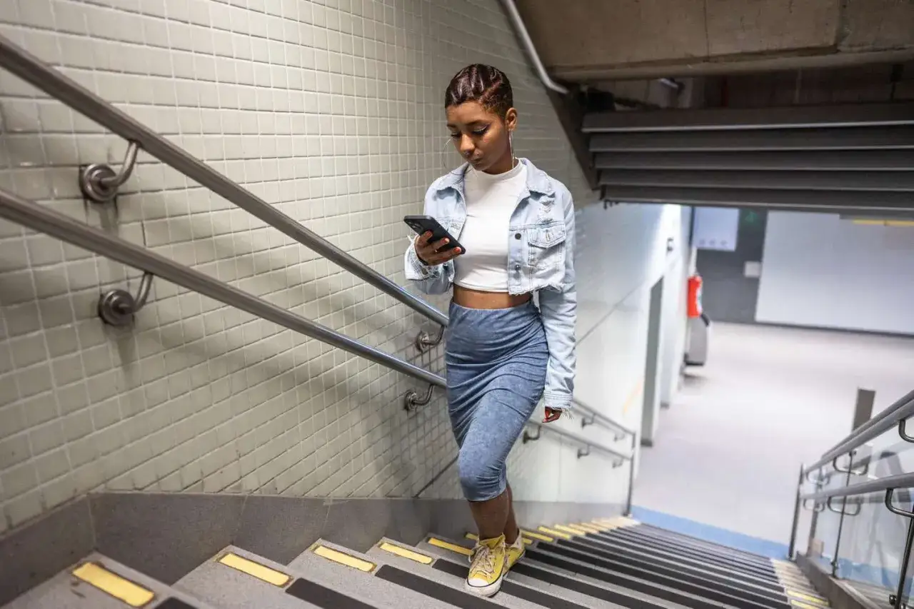 Młoda kobieta w dżinsowej kurtce i obcisłej sp&oacute;dnicy schodzi po schodach, przeglądając telefon. To nie jest bieganie po schodach, ale szybkie tempo.