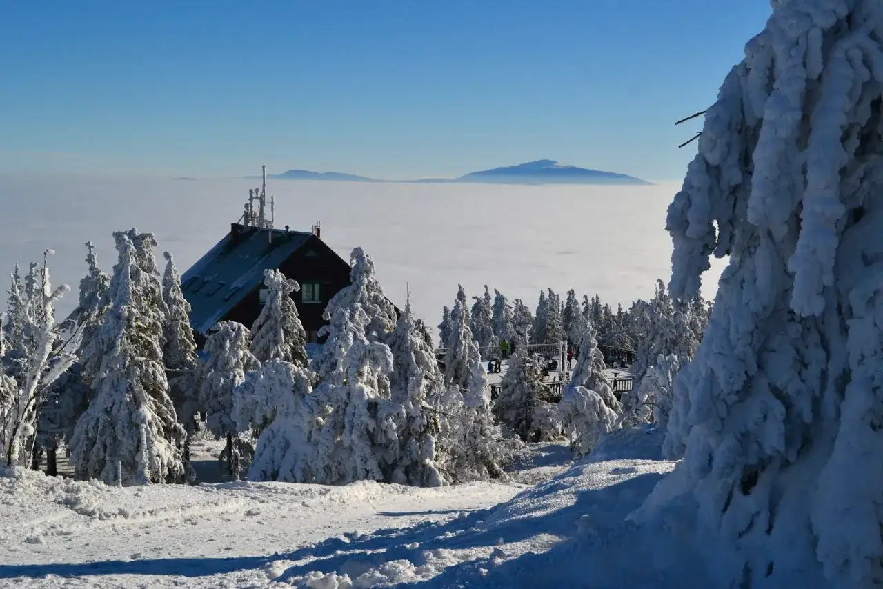 Zimowy krajobraz w Istenie. Śnieżne drzewa otaczają schronisko, a w oddali widać morze chmur i zarys g&oacute;r.