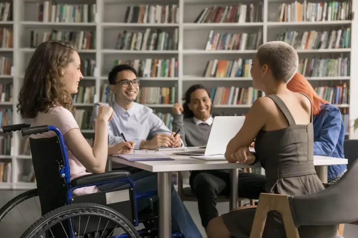 Grupa studentów dyskutuje przy stole w bibliotece. Jedna z osób porusza się na wózku, co nie stanowi przeszkody w jej nauce, mimo umiarkowanego stopnia niepełnosprawności.