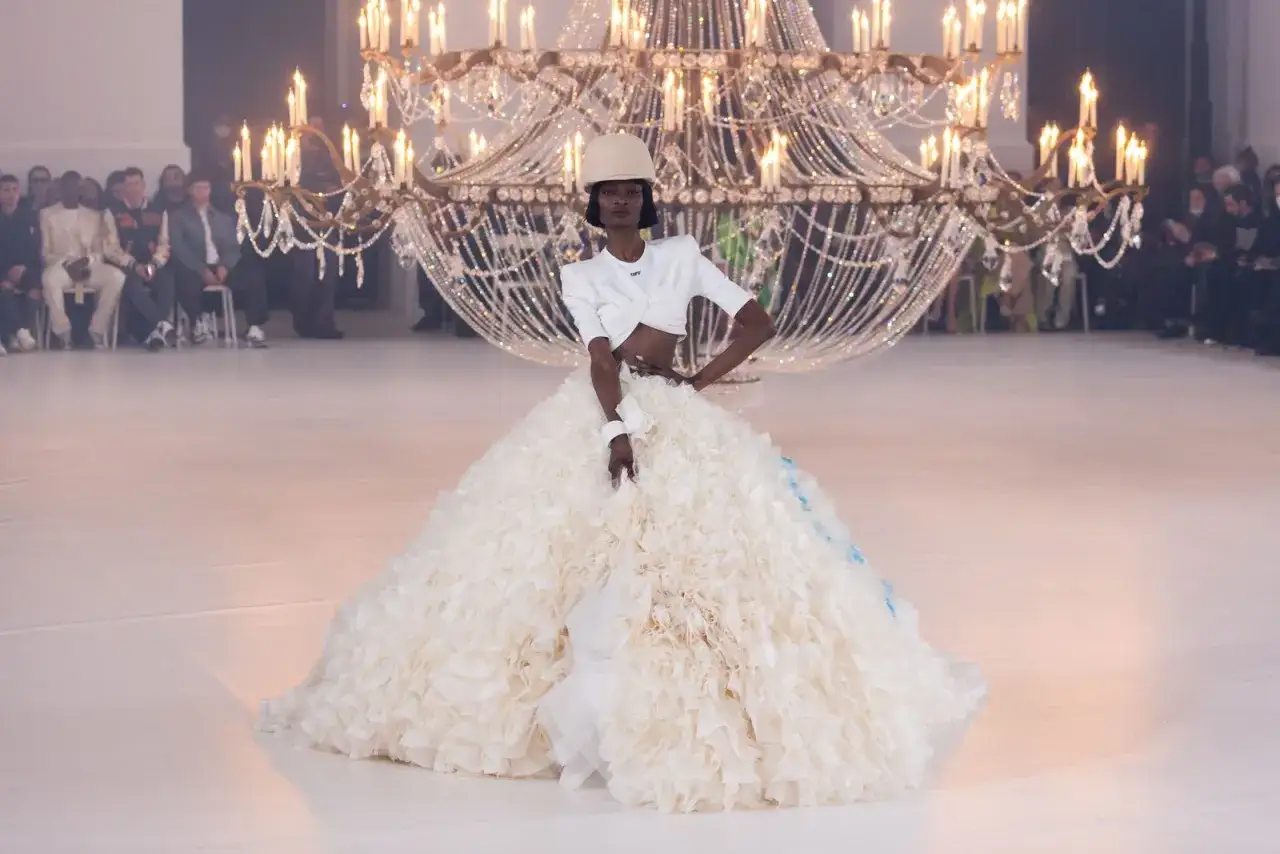 Modelka w kreacji od Off-White Virgil Abloh, z bufiastą, falbaniastą spódnicą i krótkim topem, prezentuje się pod kryształowym żyrandolem.