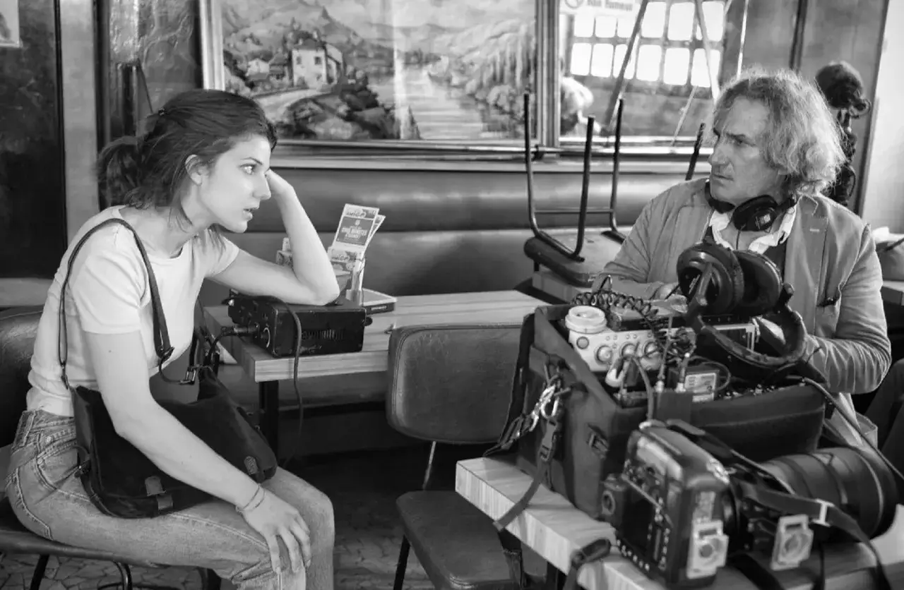 Kobieta i mężczyzna przy stoliku, jak z francuskiej nowej fali, otoczeni sprzętem filmowym.