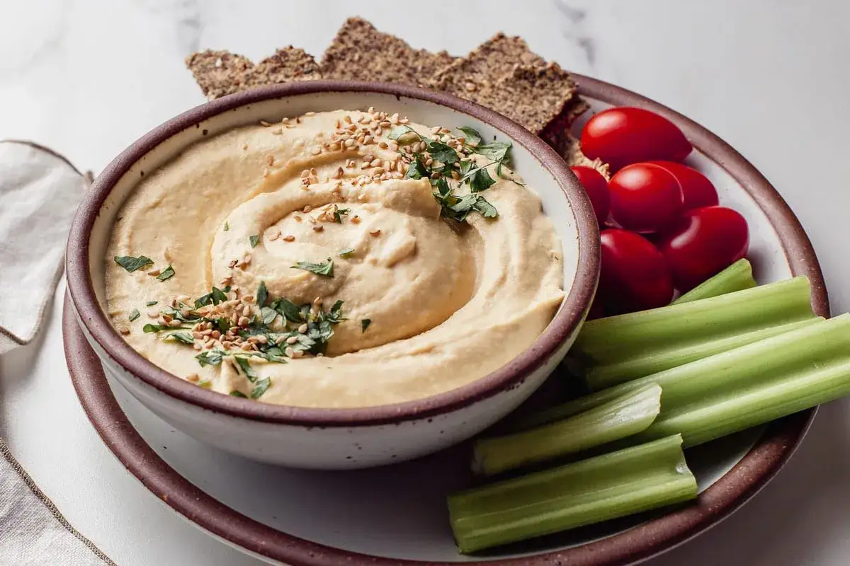 Kremowy hummus z sezamem i pietruszką, podany z pomidorkami koktajlowymi, łodygami selera i chrupkami. Idealny na zdrową przekąskę, pełną wartości odżywczych.