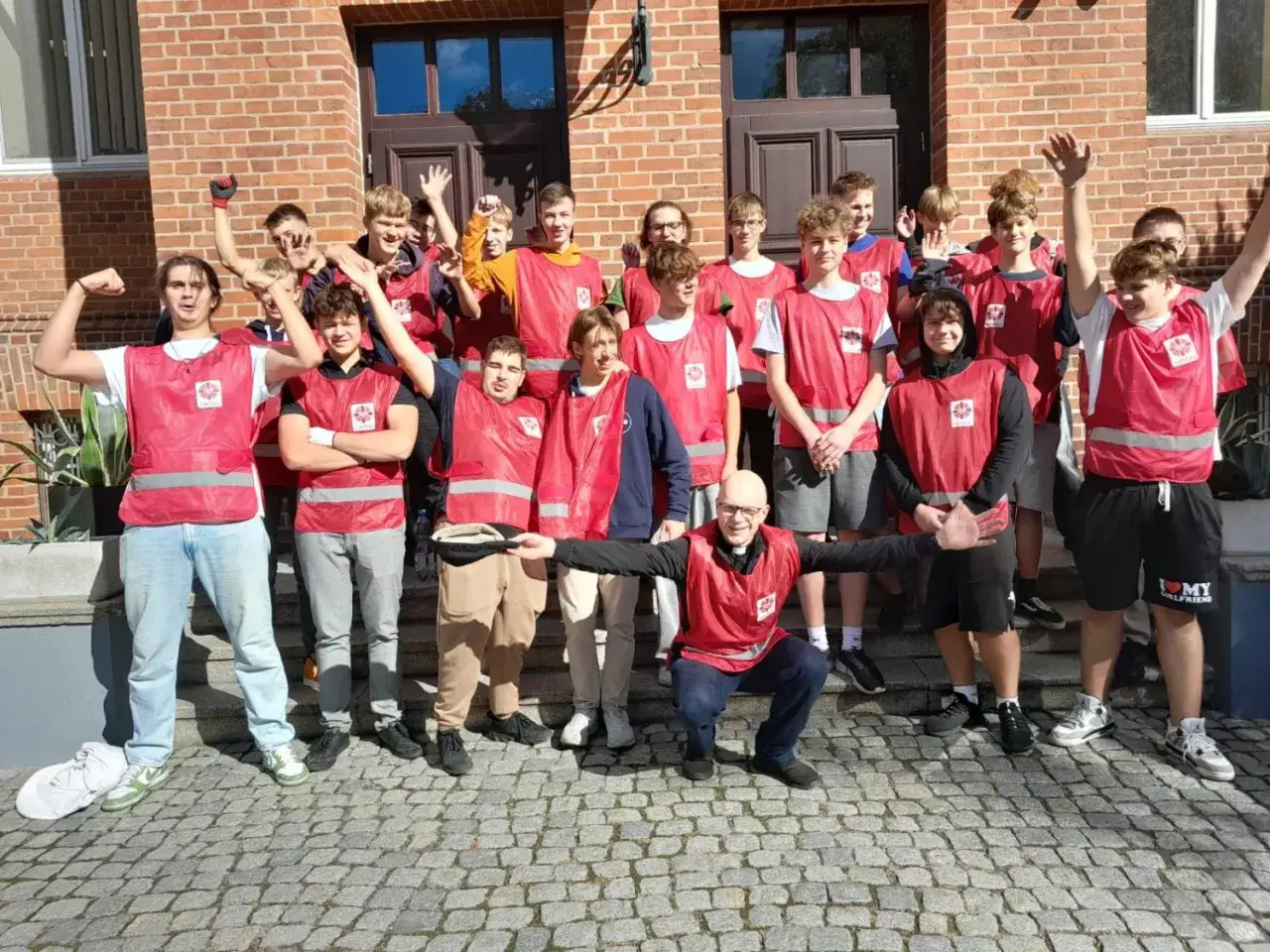 Młodzi wolontariusze w kamizelkach Caritas, w tym ksiądz, przed budynkiem centrum pomocowego Caritas im. Jana Pawła II.