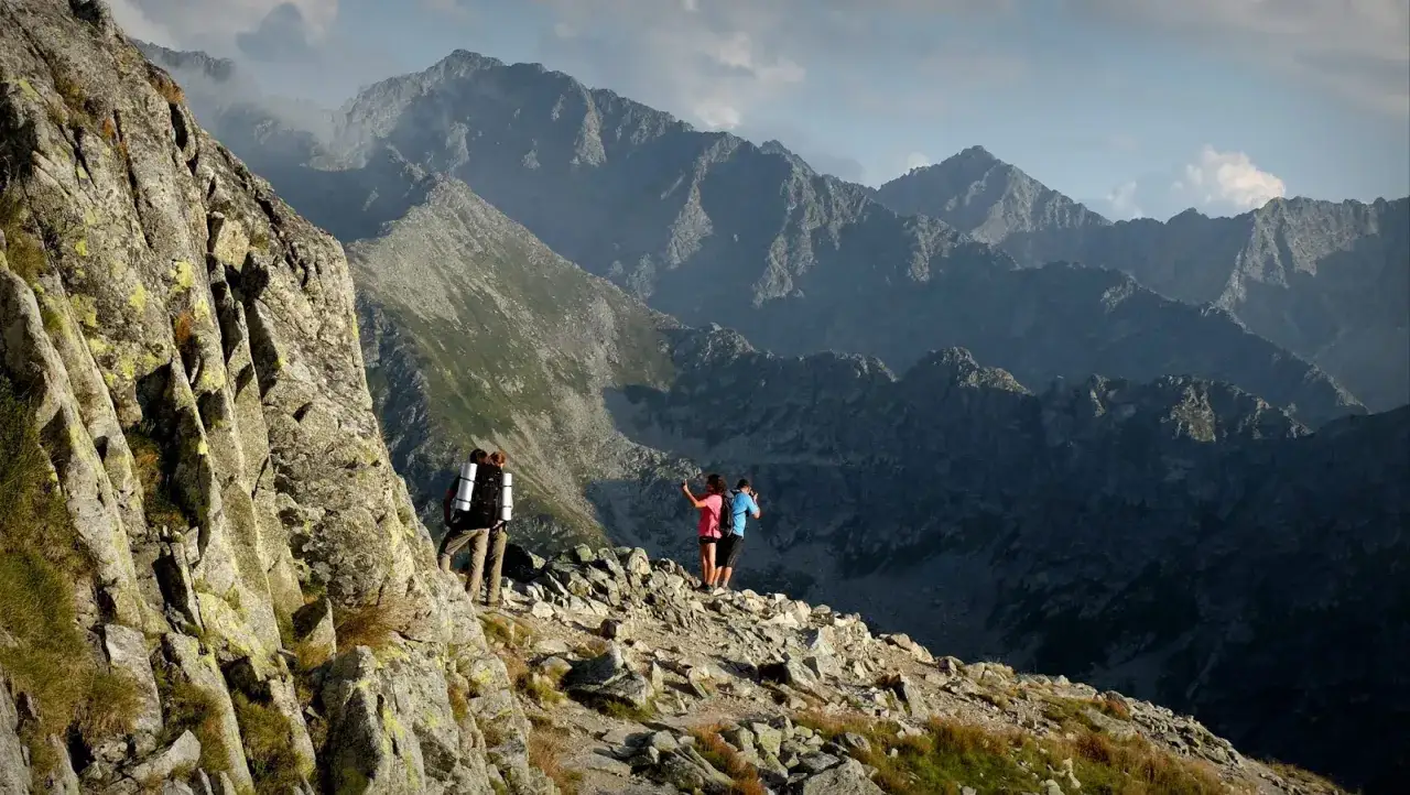 Dwie osoby robią selfie na tle majestatycznych szczytów w Tatrach. Trzecia osoba z plecakiem idzie po skalistej ścieżce.